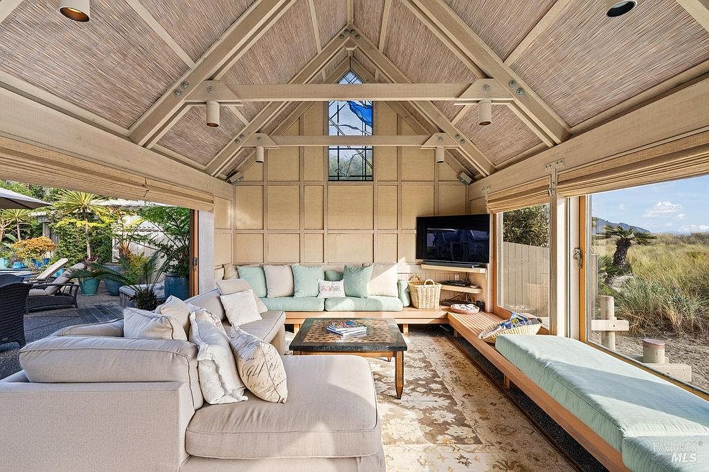 This bright and airy sunroom features a vaulted ceiling with exposed beams and a textured, woven-style ceiling finish. The space is furnished with a large, light-colored sectional sofa and a built-in bench seat that runs along the windowed wall, offering panoramic views of the outdoor landscape. A central coffee table sits on a patterned area rug, while the room opens up to an outdoor patio area, creating a seamless transition between indoor and outdoor living.