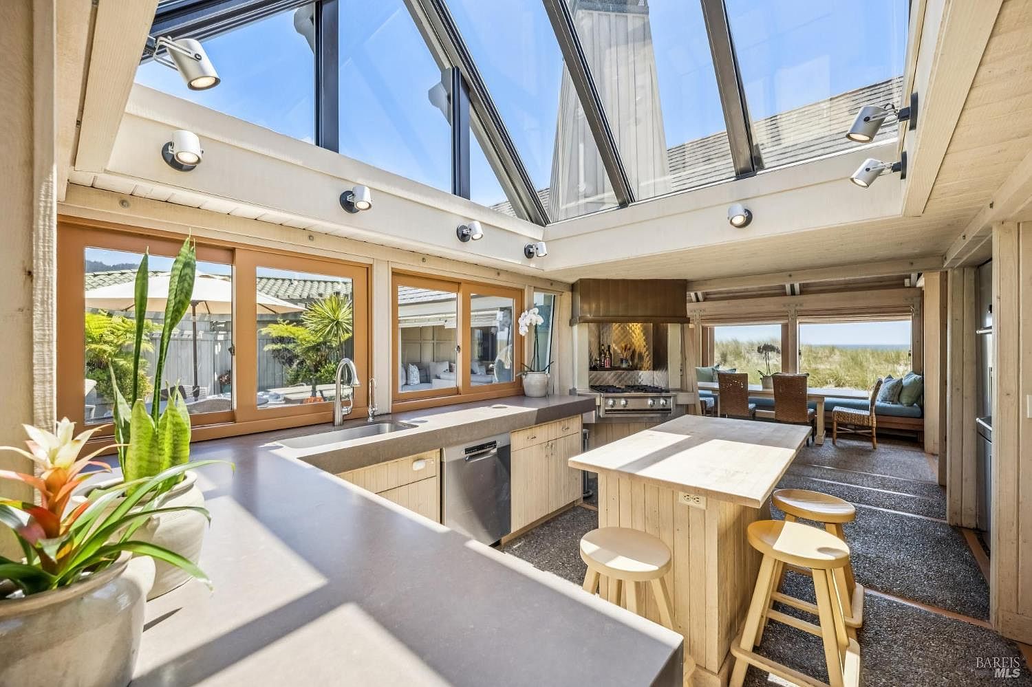 This bright, airy kitchen features a unique glass-paneled roof that floods the space with natural light, creating a conservatory-like atmosphere. The layout includes a central island with bar seating, light wood cabinetry, and a stainless steel dishwasher, all set against a backdrop of large windows that offer views of the coastal landscape. The design seamlessly blends indoor cooking space with the surrounding outdoor environment, emphasizing an open and relaxed aesthetic.