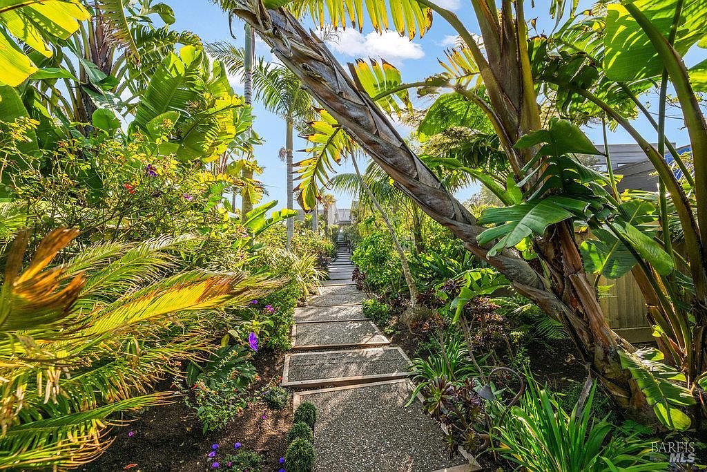 A lush, tropical-inspired garden path leads through a dense collection of palm trees, banana plants, and various ornamental shrubs. The walkway is constructed from rectangular stone pavers set into a gravel base, creating a serene and private atmosphere. The perspective is a low-angle, eye-level shot looking down the path, emphasizing the vibrant greenery and the dappled sunlight filtering through the canopy.
