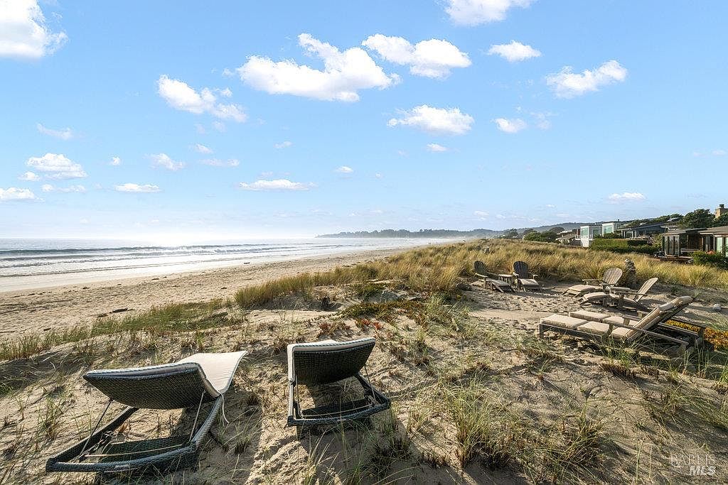 This scenic outdoor space features a private sandy dune area equipped with multiple lounge chairs, offering direct access to a vast, tranquil beach. The perspective is captured from a slightly elevated position on the dunes, looking out toward the horizon where the ocean meets a clear blue sky with scattered clouds. The setting provides a serene and luxurious coastal atmosphere, highlighting the property's prime beachfront location.