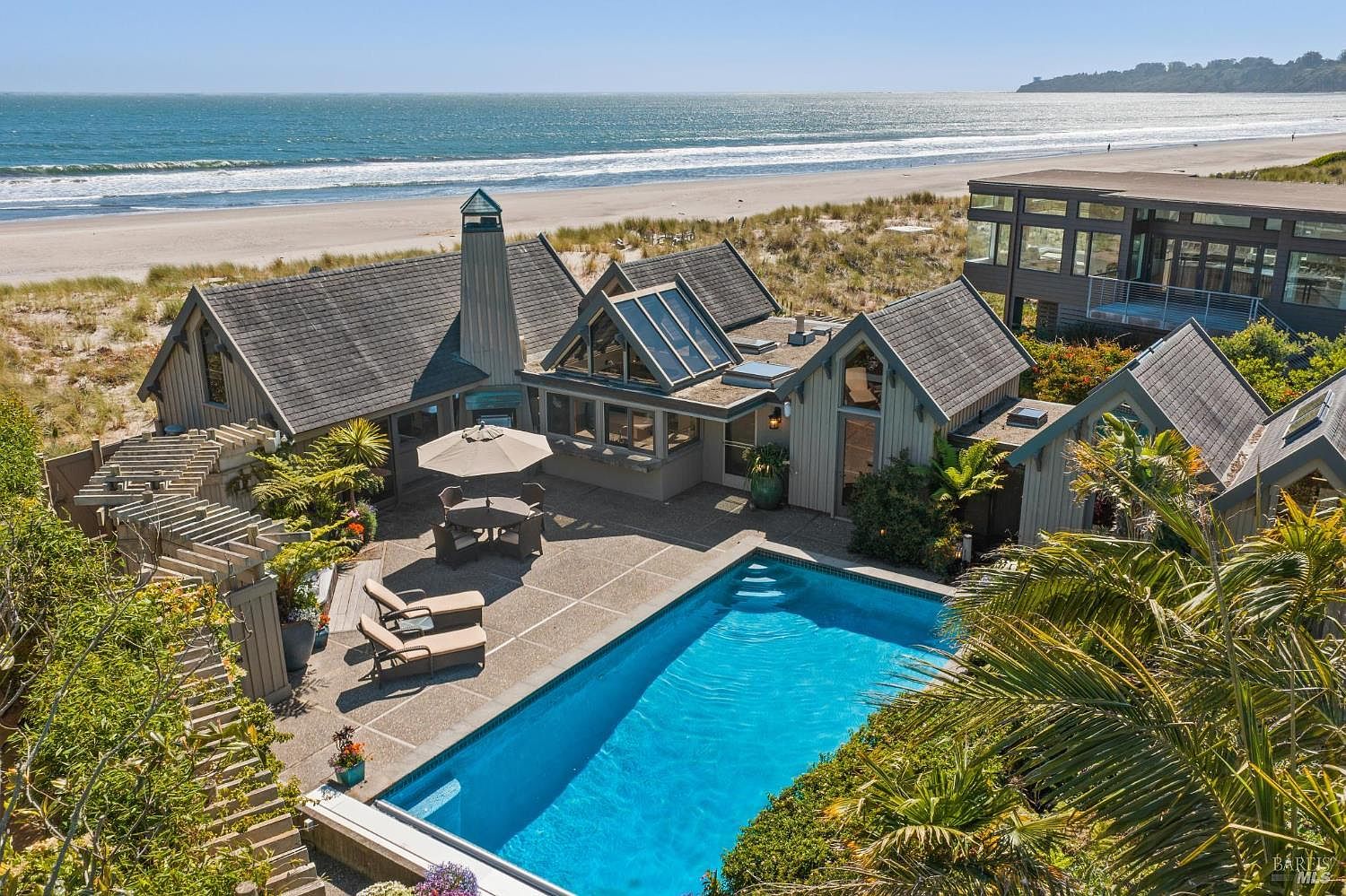 This high-angle aerial shot captures a coastal residence featuring a multi-gabled roofline and a private swimming pool set within a paved patio area. The property is situated directly adjacent to a sandy beach and dunes, offering expansive ocean views. A secondary structure with large windows is visible to the right, and the overall aesthetic is that of a secluded, upscale beach retreat.