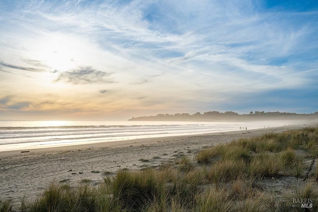 This scenic view captures a wide, sandy beach at sunset, highlighting the natural beauty of the coastal landscape. The foreground features tall, wind-swept beach grasses, while the gentle surf rolls onto the shore under a soft, hazy sky. This image emphasizes the tranquil, expansive outdoor lifestyle and direct beach access available to the local community.