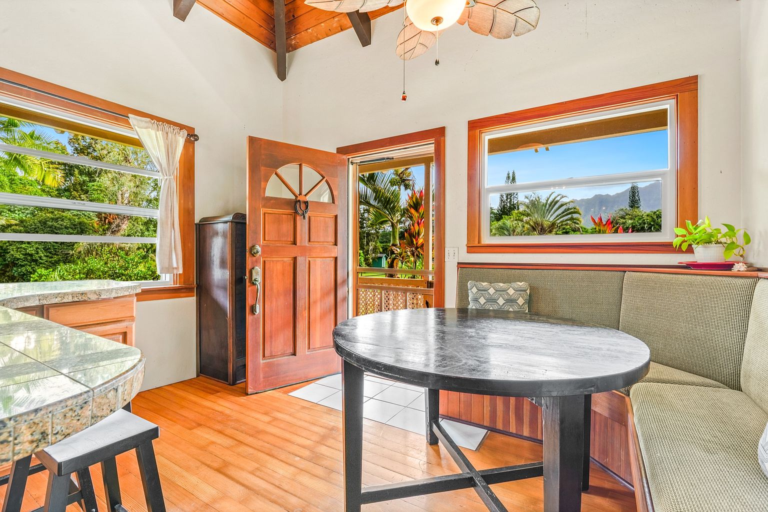 This dining room features a round, dark wood table with seating that includes a built-in bench and a stool. Natural light floods the space through two windows, offering views of lush greenery and a mountain range. The room's design incorporates warm wood tones and a cozy, inviting atmosphere.