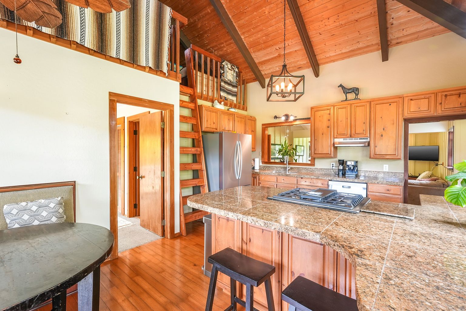 This is a warm and inviting kitchen with wood cabinetry, granite countertops, and stainless steel appliances. The kitchen features a central island with bar seating and a gas cooktop. A wooden ladder leads to a loft area, adding a unique architectural element to the space, and the hardwood floors contribute to the cozy atmosphere.