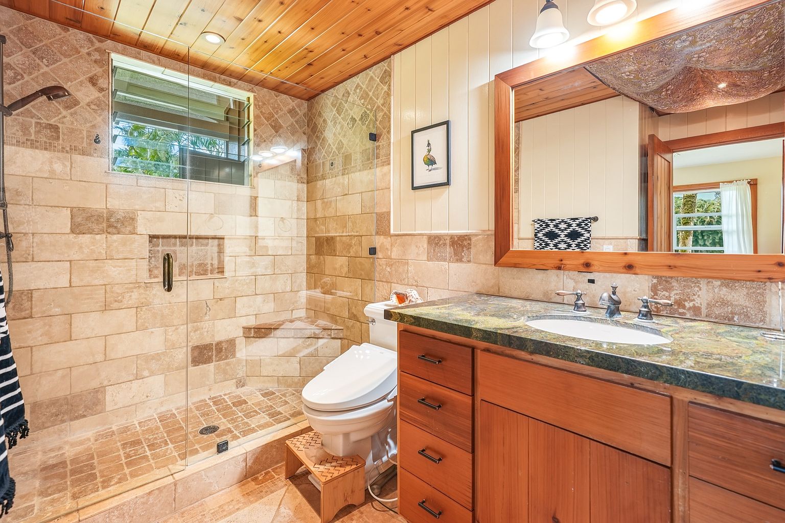 This is a primary bathroom featuring a walk-in shower with glass doors and tiled walls, a toilet with a wooden step stool, and a wooden vanity with a green marble countertop. The bathroom has a warm, natural aesthetic with wood paneling on the ceiling and walls, complemented by the earth-toned tiles. A framed mirror hangs above the sink, reflecting the natural light from the window.
