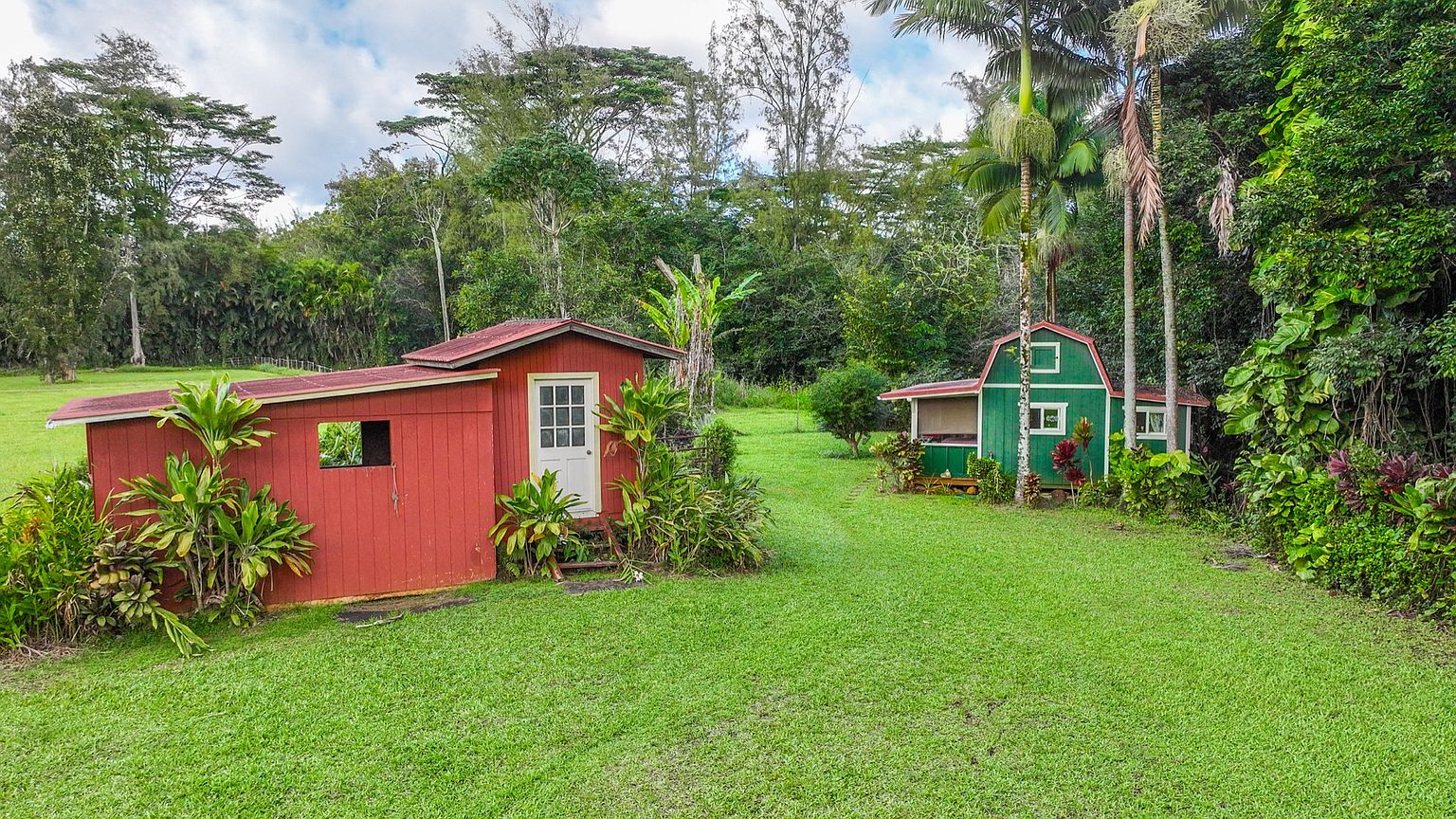 This image showcases a lush yard with two small outbuildings. The building on the left is painted red with a white door, while the one on the right is green with a barn-style roof. The vibrant green lawn and surrounding tropical foliage create a serene and inviting atmosphere, suggesting a peaceful and private property.