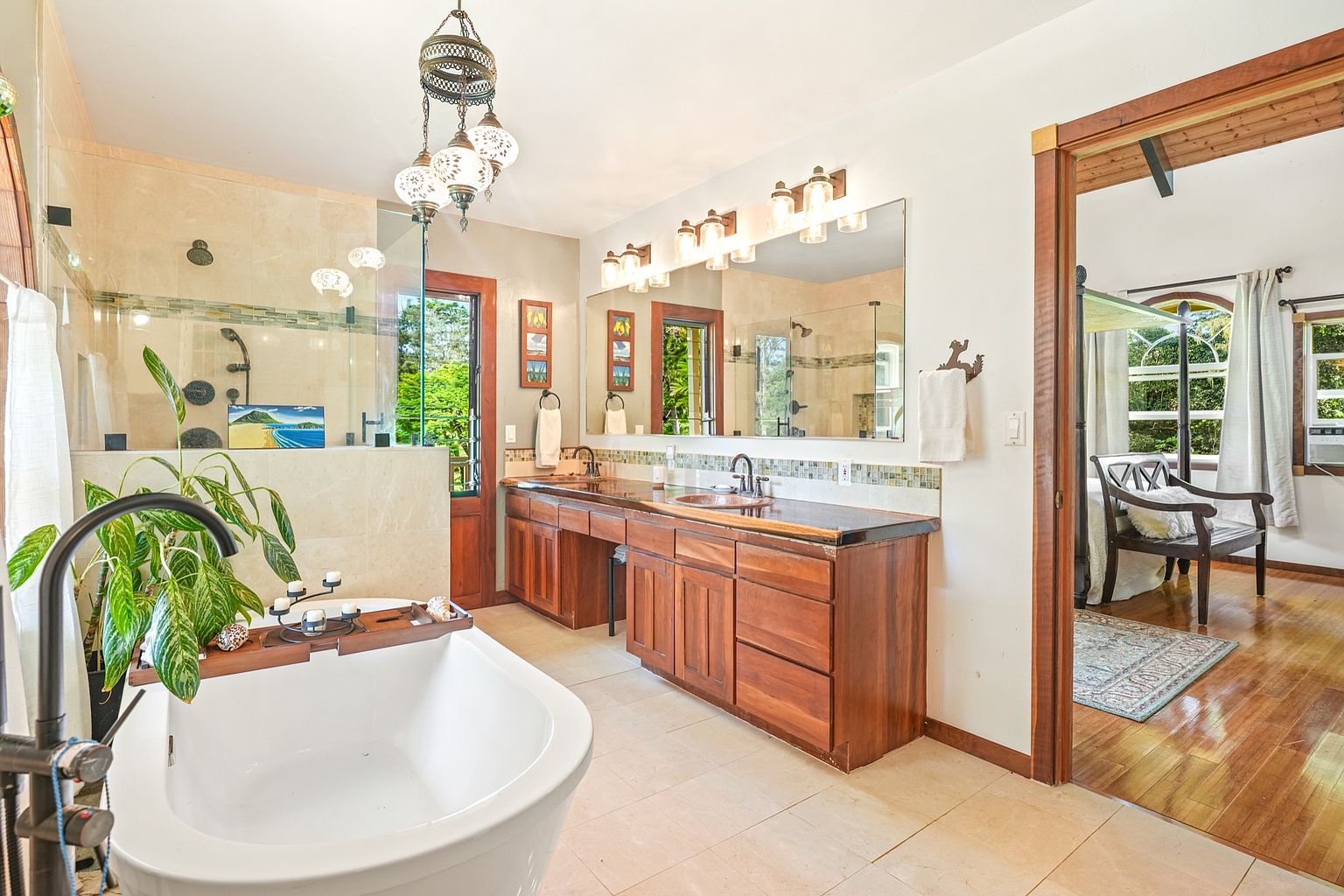 This is a primary bathroom featuring a freestanding bathtub with a black faucet, a wooden vanity with dual sinks and a large mirror, and a glass-enclosed shower. The room is decorated with plants and decorative lighting, creating a spa-like atmosphere. The style is luxurious and inviting, with warm wood tones and natural light.