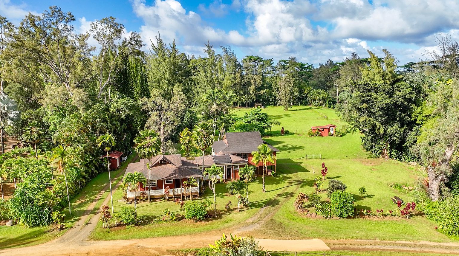 This aerial shot showcases a sprawling property featuring a large, multi-sectioned red house with dark roofing, surrounded by lush greenery, including palm trees and dense forest. A well-maintained lawn extends from the house, leading to a smaller red outbuilding in the distance, with a dirt driveway providing access to the property. The overall impression is one of a secluded, private estate with ample outdoor space.