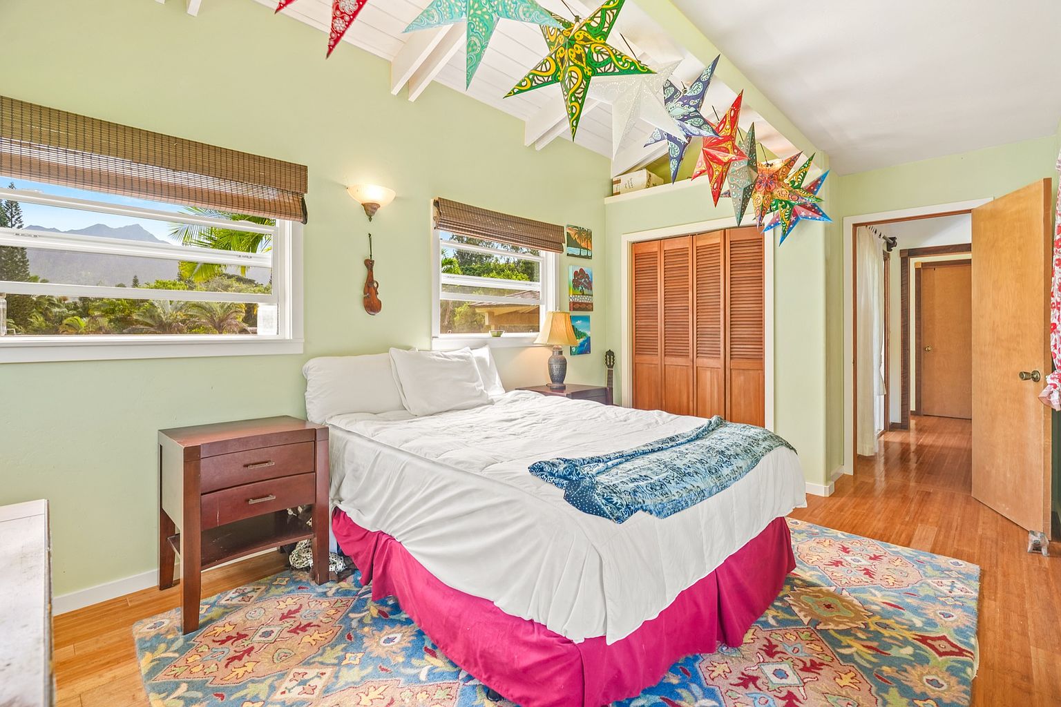 This is a cozy bedroom featuring light green walls, a bed with white linens and a blue throw, and a colorful area rug. The room has two windows with bamboo blinds, a wooden nightstand, and a closet with louvered doors. Decorative star lanterns hang from the ceiling, adding a whimsical touch to the space.