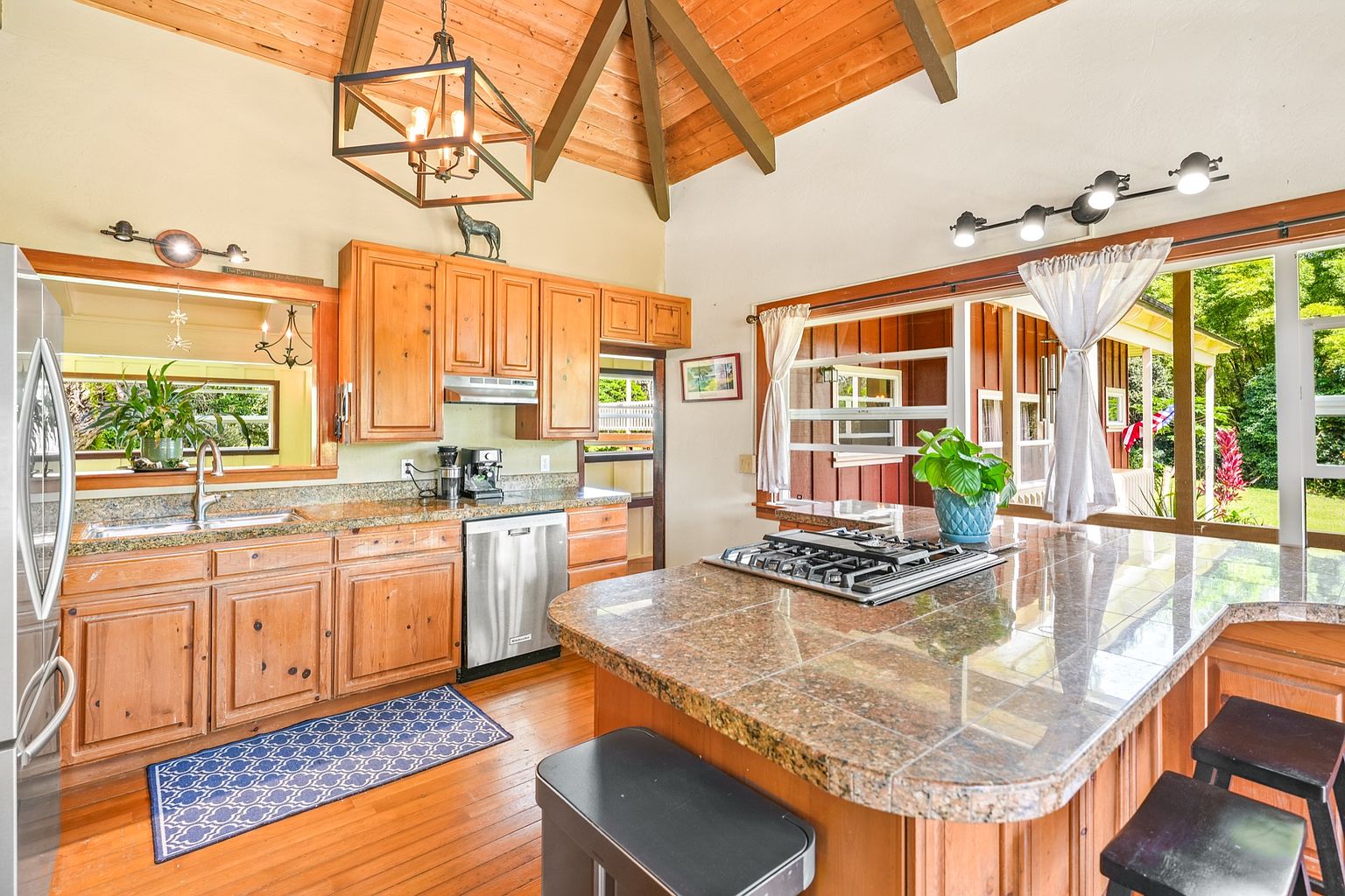 This is a warm and inviting kitchen featuring wooden cabinets, granite countertops, and stainless steel appliances. A central island with a cooktop and seating provides a focal point, while large windows offer views of the outdoors. The vaulted ceiling with exposed beams adds a rustic touch to the space.