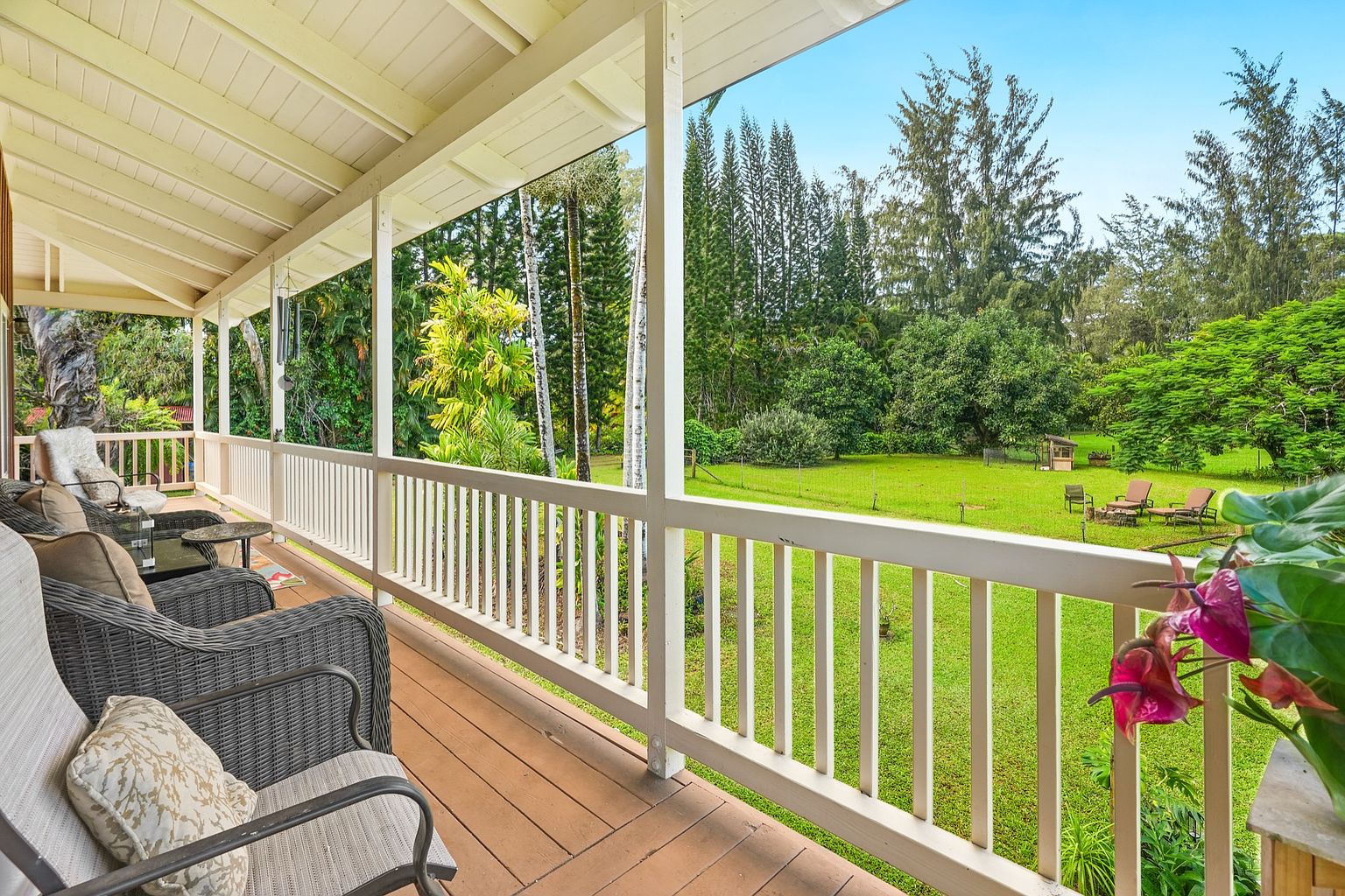This image showcases a charming patio or deck area, featuring comfortable wicker furniture and a wooden floor. The space is framed by a white railing, offering an inviting view of a lush green lawn and mature trees. The scene evokes a sense of relaxation and outdoor living.