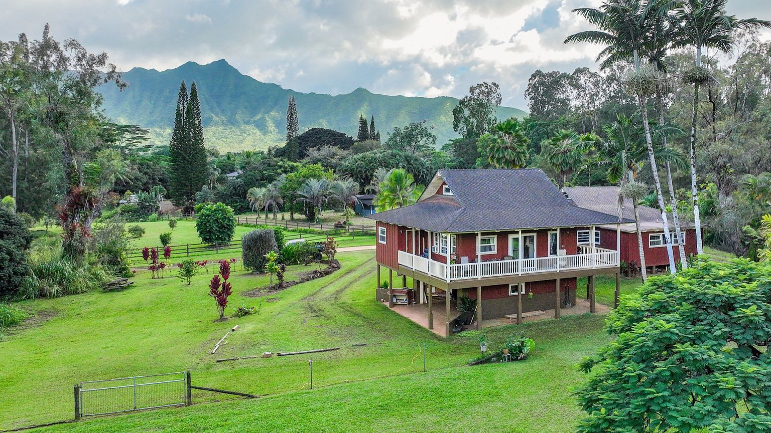 This aerial view showcases a charming, elevated red house with a dark roof, surrounded by lush greenery and a well-maintained lawn. The property features a white-railed deck, adding to its appeal, and is set against a backdrop of scenic mountains and a partly cloudy sky. The overall impression is one of tranquility and natural beauty, highlighting the property's desirable location and inviting exterior.