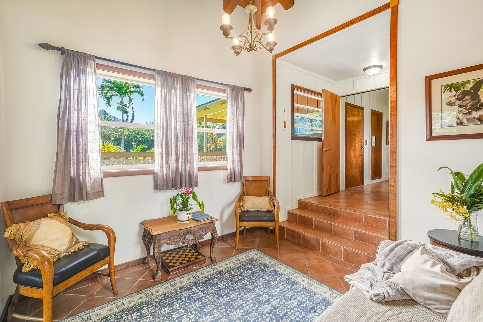 This inviting living room features a cozy seating area with a blue patterned rug, two wooden chairs, and a comfortable sofa. Natural light streams in through the windows, illuminating the room and highlighting the warm, earthy tones of the tile flooring and wooden accents. The space is decorated with plants and artwork, creating a welcoming and comfortable atmosphere.