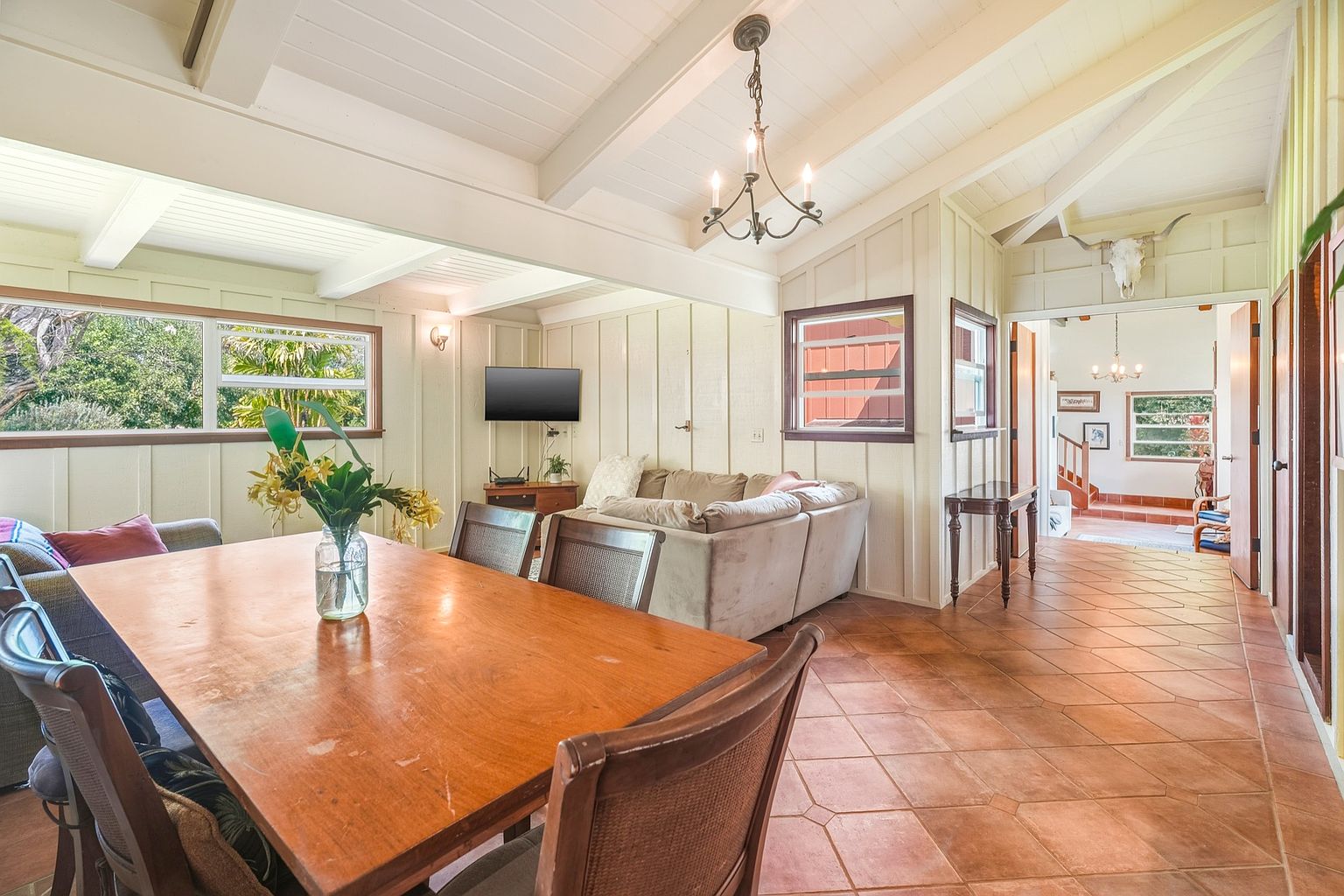 This interior shot showcases a dining room with a large wooden table and chairs, complemented by a view into the living room area. The room features a unique architectural style with white wooden beams on the ceiling and a tiled floor, creating a warm and inviting atmosphere. Natural light streams in through the windows, enhancing the room's appeal.