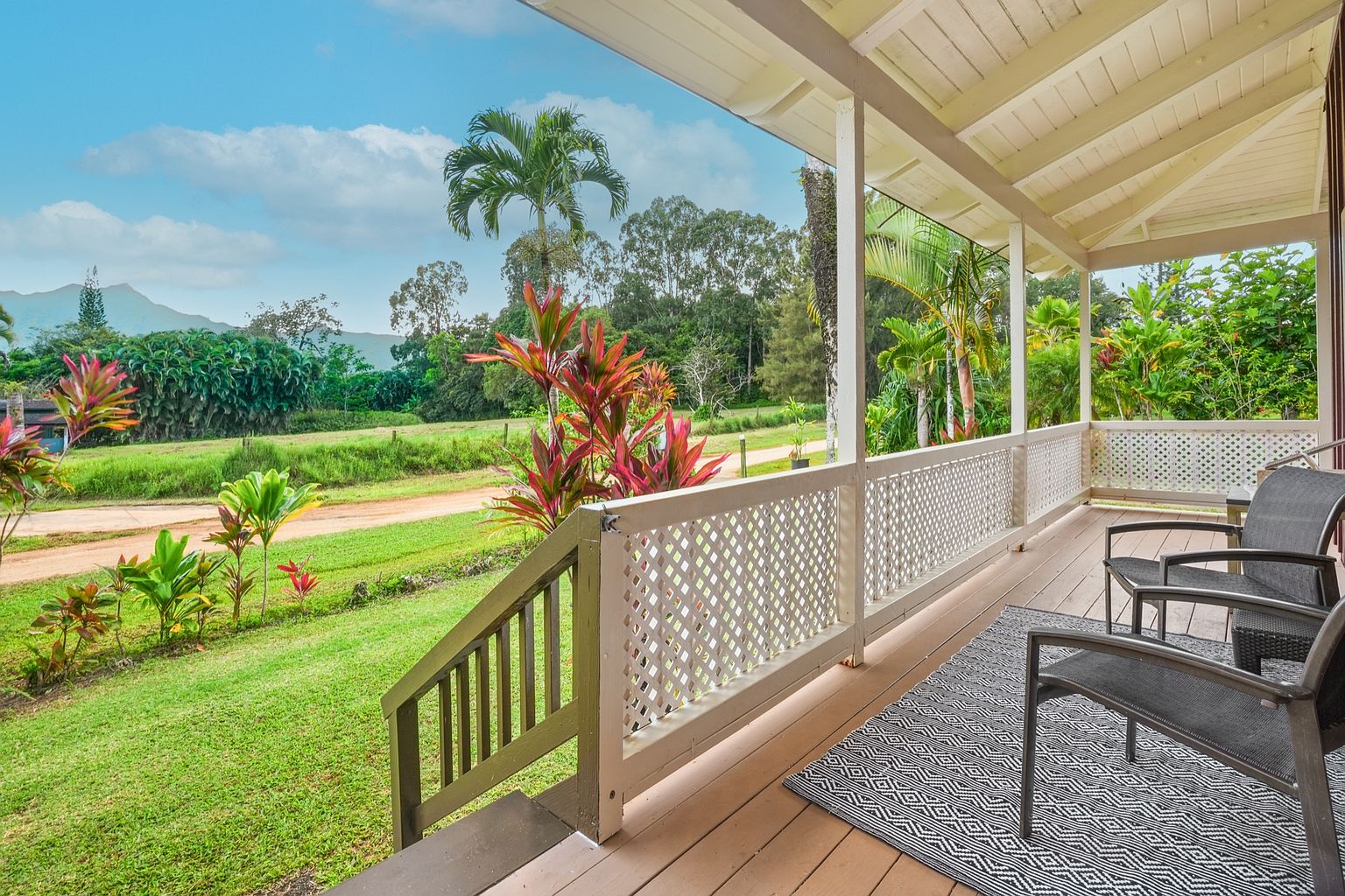 This image showcases a charming patio or deck area, featuring a wooden floor and a lattice railing. Comfortable seating is arranged on a patterned rug, inviting relaxation. The view extends to a lush green lawn and tropical foliage, creating a serene and inviting outdoor living space.