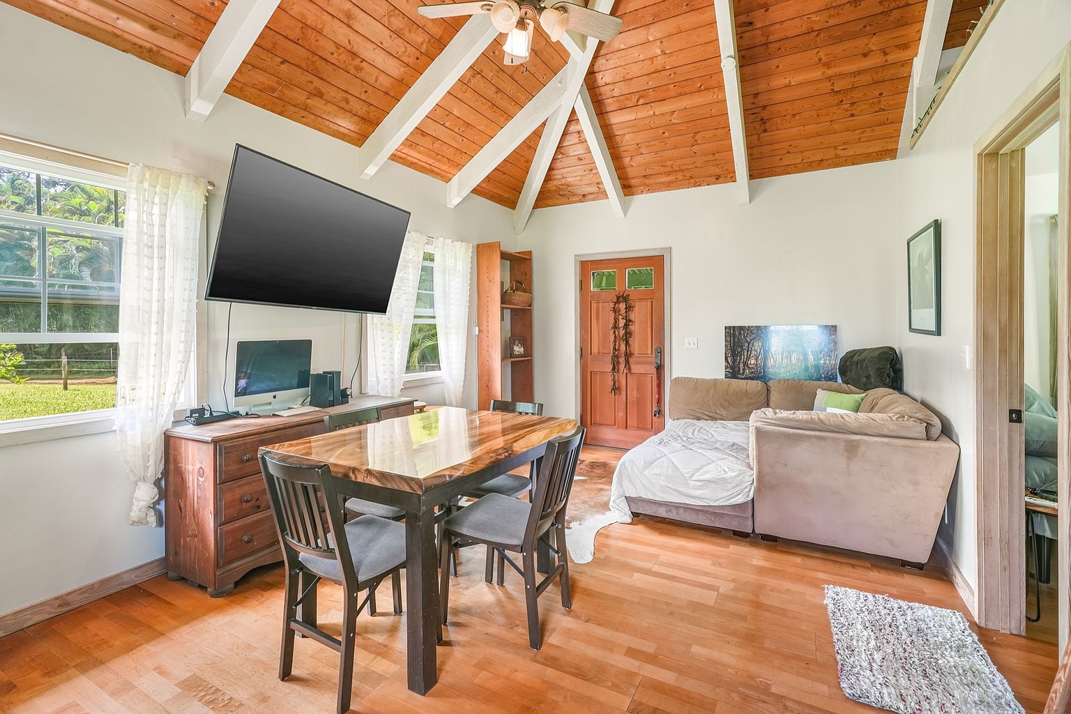 This interior shot showcases a cozy living space with a dining area. The room features a wooden ceiling with white beams, hardwood floors, and a comfortable sofa. A large television is mounted on the wall above a wooden dresser, and natural light streams in through the windows, creating a warm and inviting atmosphere.