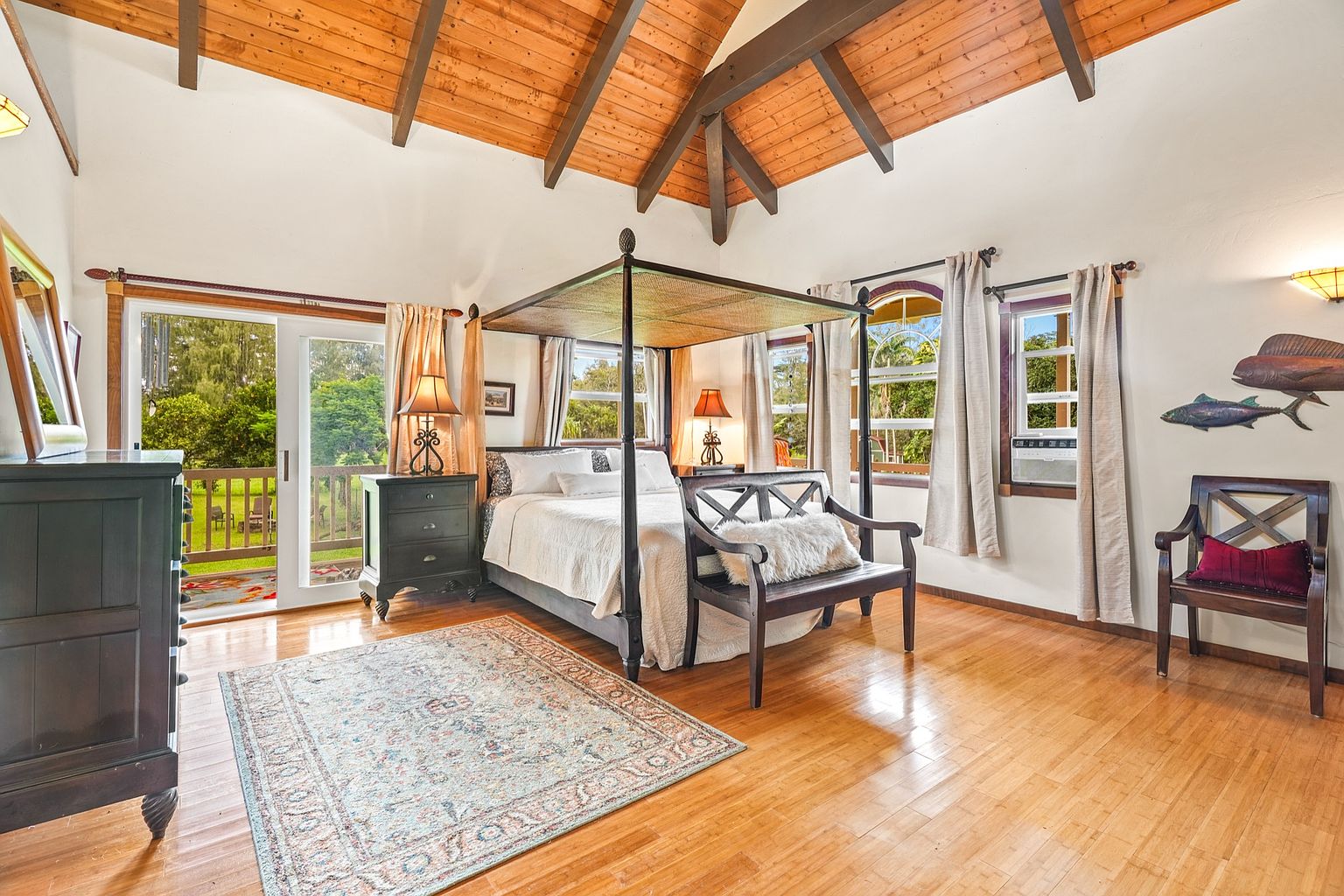This is a primary bedroom featuring a four-poster bed with a light-colored comforter and pillows. The room has hardwood floors, a vaulted wood ceiling, and multiple windows that provide natural light. There is a dark wood dresser and nightstand, as well as a bench at the foot of the bed and a chair in the corner. A rug is placed in front of the bed.