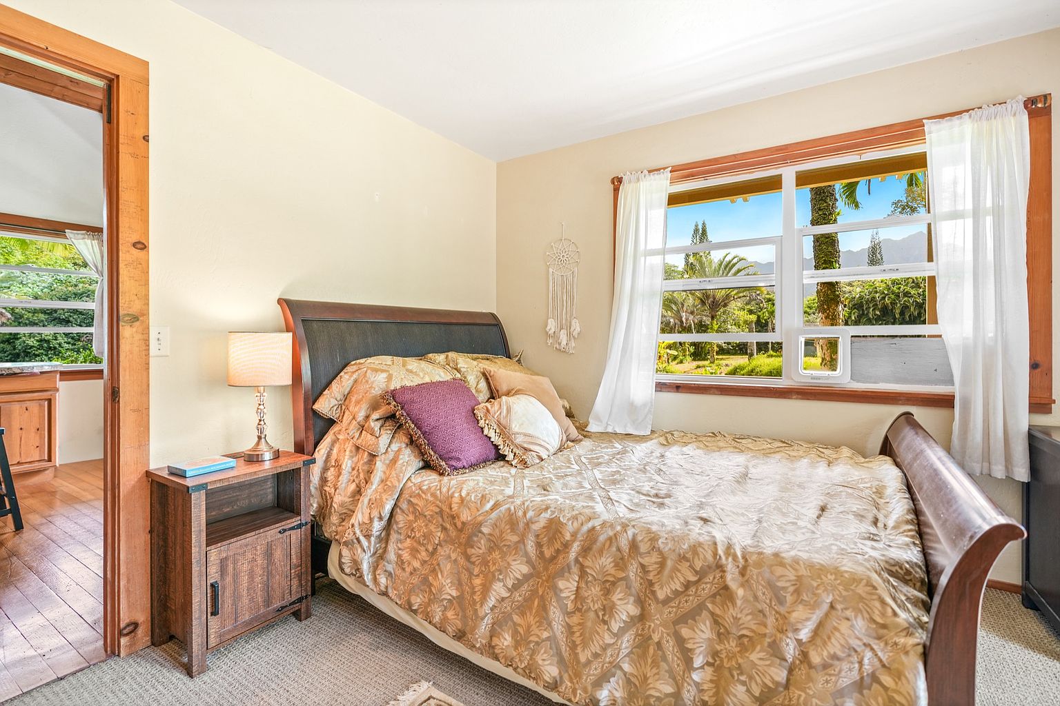 This is a cozy bedroom featuring a wooden-framed bed with a patterned gold comforter and decorative pillows. A wooden nightstand with a lamp sits beside the bed, and a window with white curtains offers a view of lush greenery outside. The room has a warm and inviting atmosphere, perfect for a guest room.