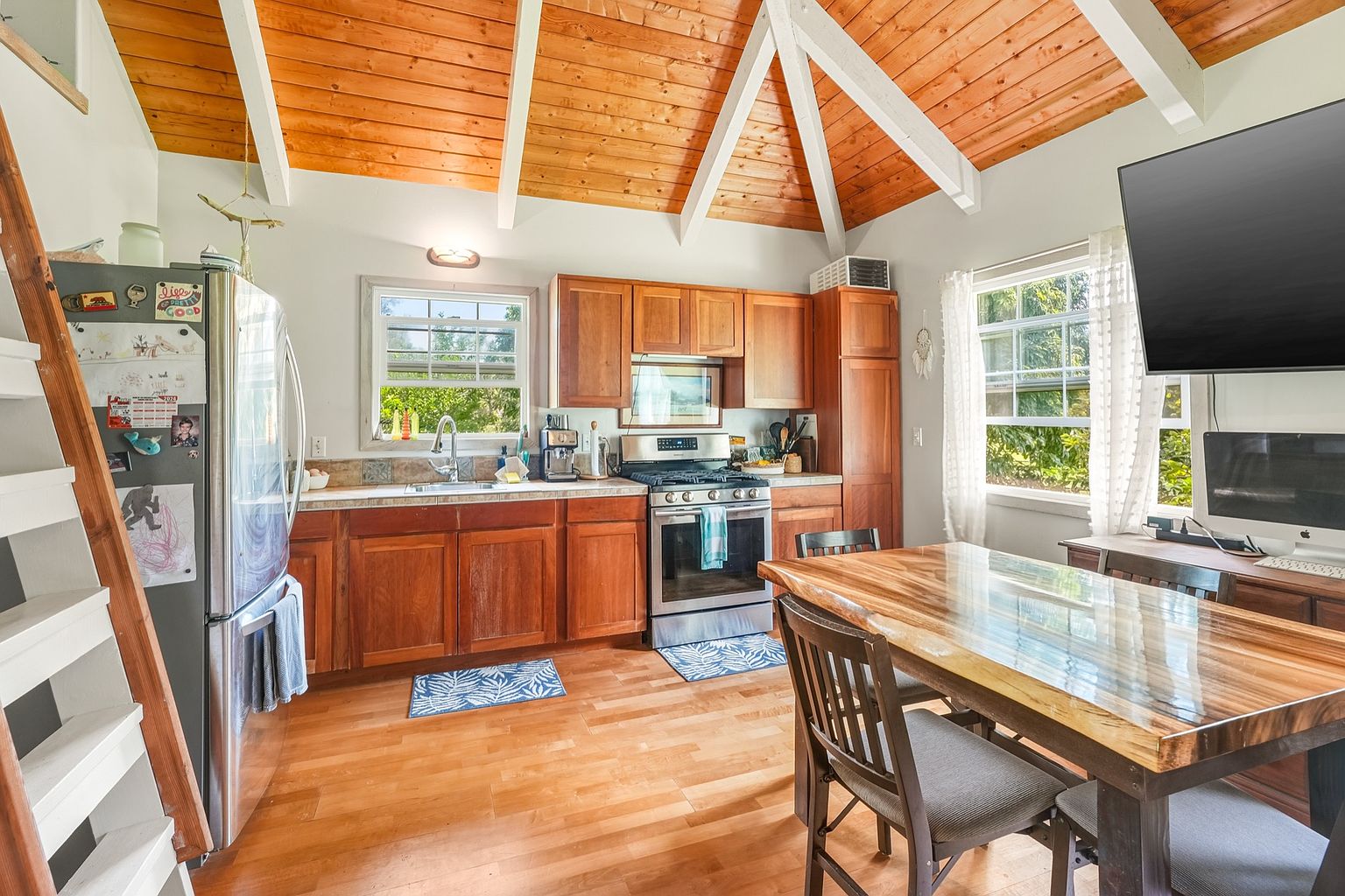 This is a warm and inviting kitchen featuring wooden cabinets, stainless steel appliances, and a unique wooden ceiling with white beams. A wooden dining table sits adjacent to the kitchen area, creating a cozy and functional space. The room is well-lit by natural light streaming through the windows, enhancing the overall appeal.