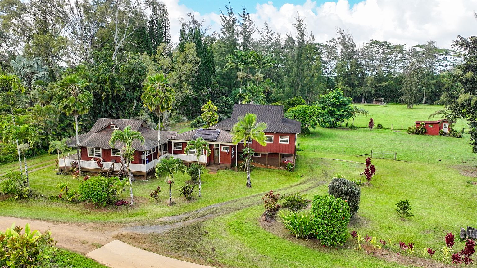This aerial shot showcases a charming red house nestled amidst lush greenery and tall trees. The property features a well-maintained lawn, a winding driveway, and a separate small red outbuilding in the distance. The overall impression is one of serene country living with a touch of rustic elegance.
