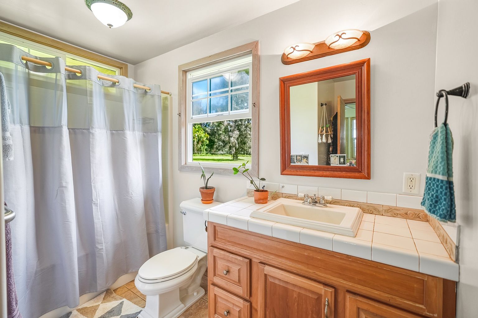 This is a well-lit bathroom featuring a wooden vanity with a white tiled countertop and sink. A framed mirror hangs above the sink, complemented by a light fixture. A toilet is positioned next to a window, and a shower with a curtain is visible on the left, creating a functional and inviting space.