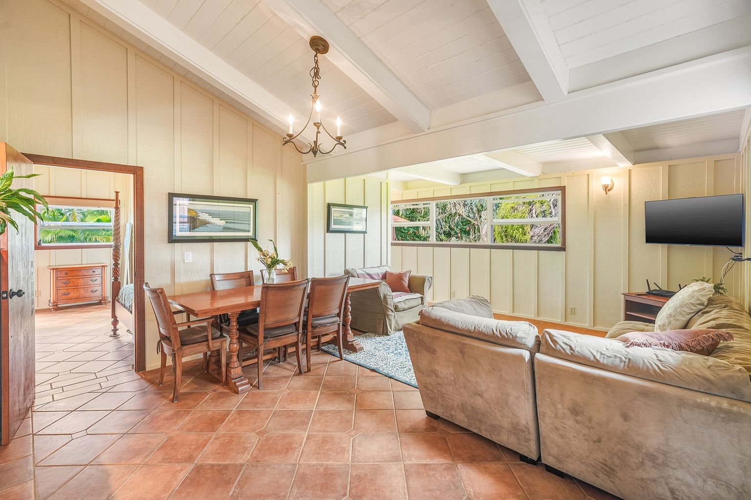 This interior shot showcases a combined living and dining area with a warm, inviting ambiance. The room features terracotta tile flooring, light yellow paneled walls, and a white beamed ceiling. A wooden dining table with chairs sits adjacent to a comfortable living area with a large sofa and armchair, creating a cozy and functional space.