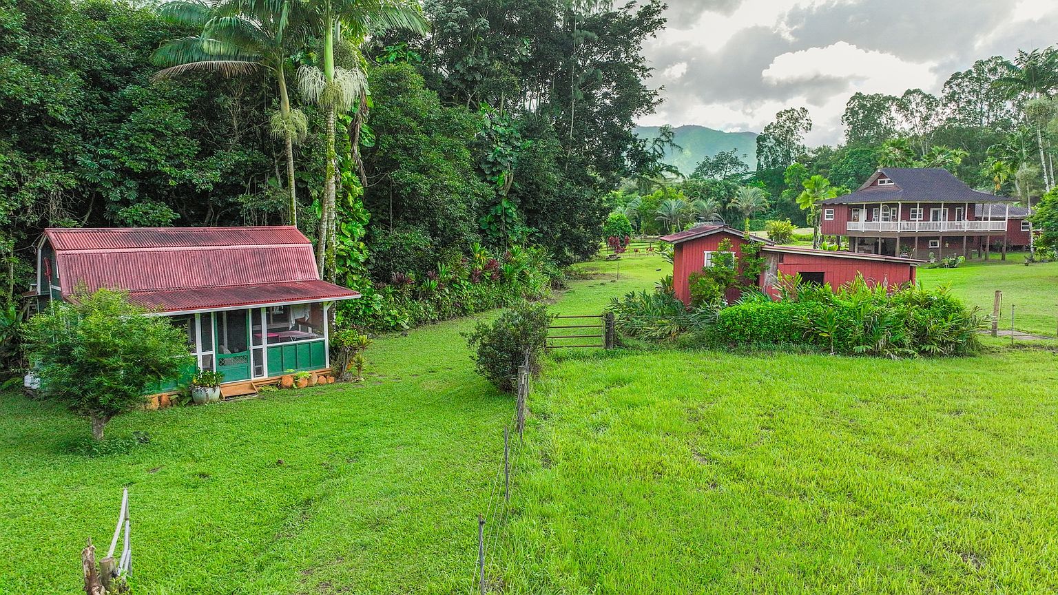 This aerial view showcases a charming property with lush green lawns and mature trees. A quaint cottage with a red roof and green trim sits to the left, while a larger two-story house with a balcony is visible in the background, along with a red outbuilding. The landscape is well-maintained, creating a serene and inviting atmosphere.