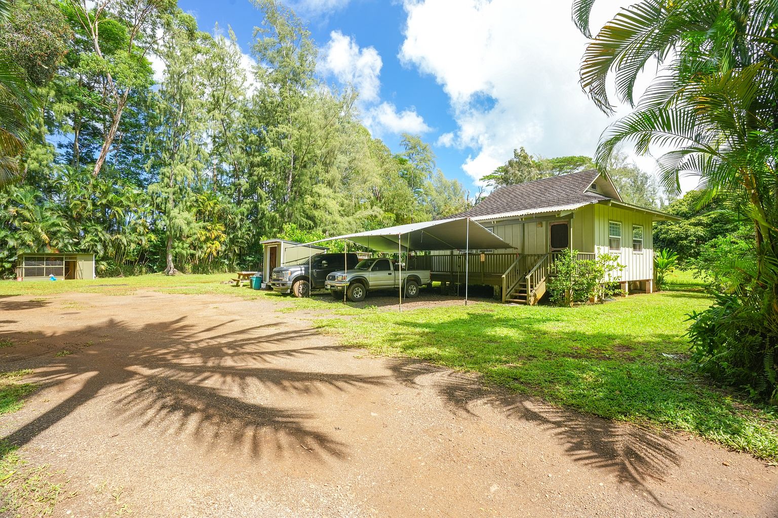 This image showcases the front exterior of a charming home with a well-maintained yard. The house features a covered parking area, a small porch with stairs, and light-colored siding. Lush greenery surrounds the property, creating a serene and inviting atmosphere.