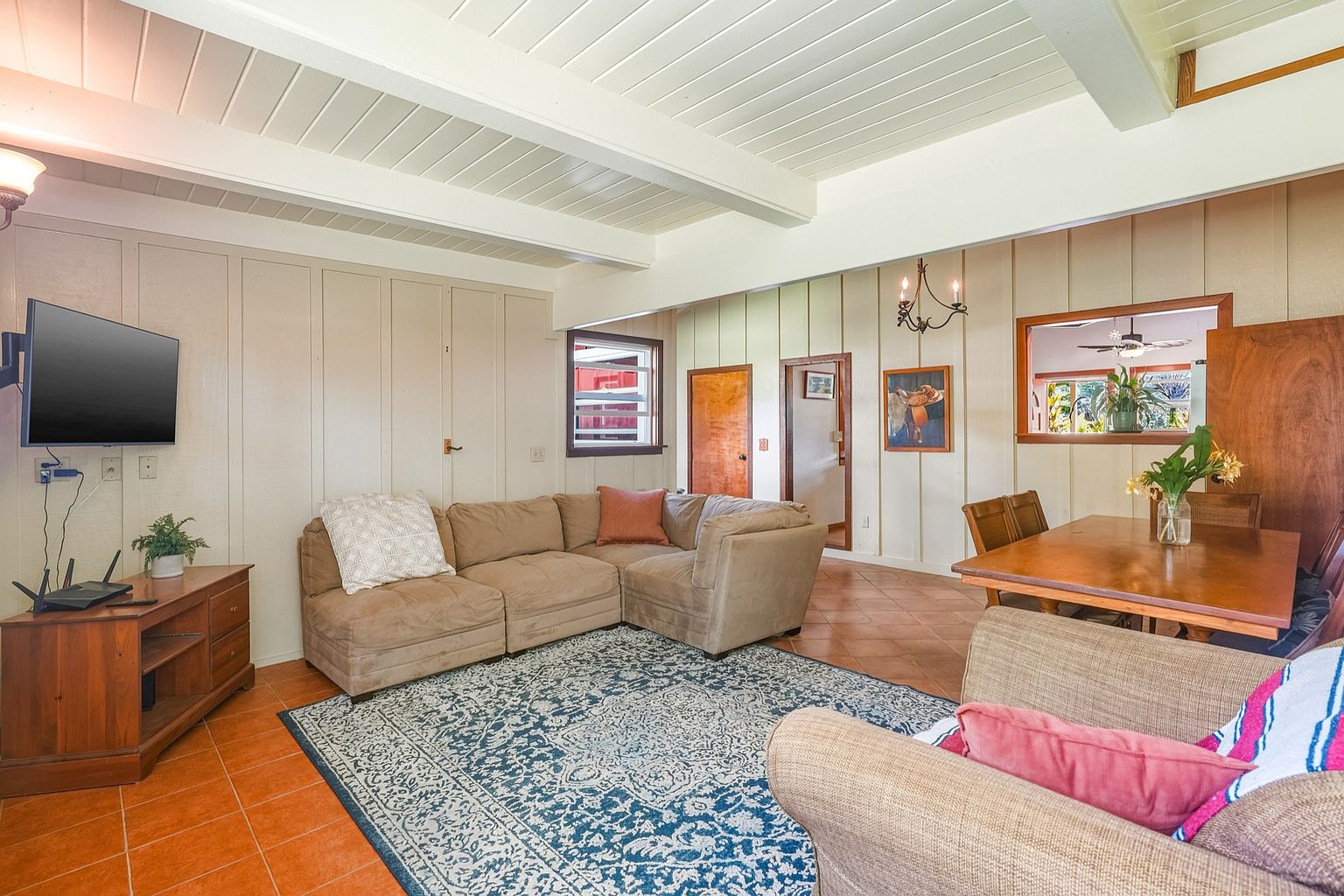 This inviting living room features a comfortable sectional sofa arranged on a patterned blue rug, creating a cozy gathering space. A wooden entertainment center houses a television and other electronics, while the adjacent dining area includes a wooden table and chairs. The room is well-lit and has a relaxed, homey atmosphere.