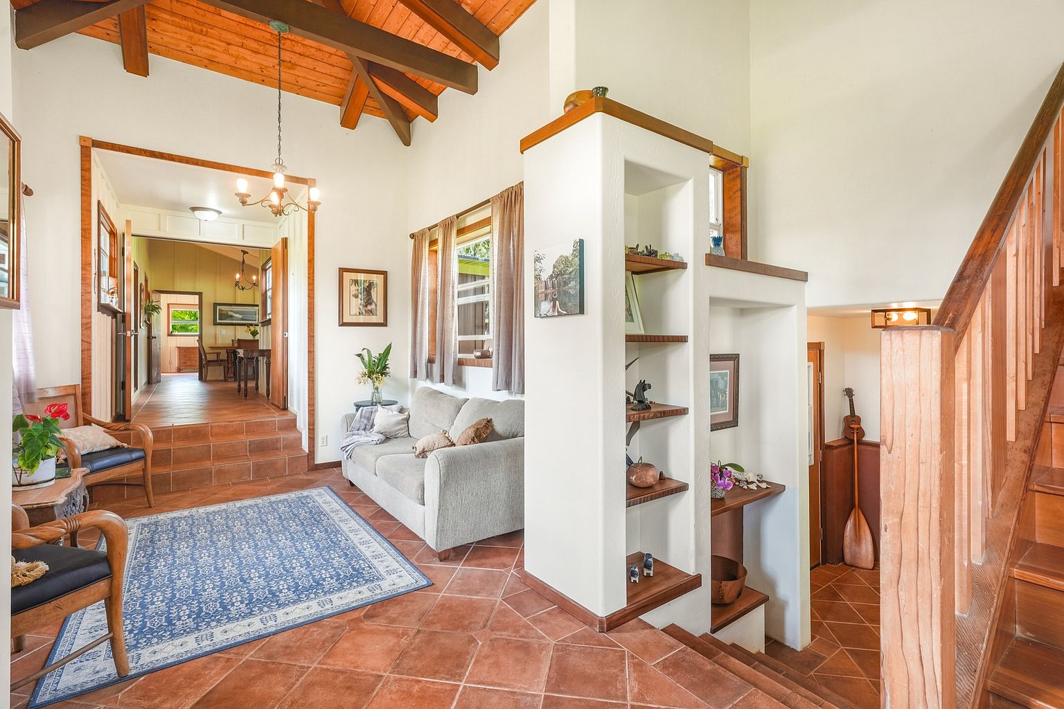 This inviting living room features terracotta tile flooring, a blue patterned rug, and a comfortable gray sofa. The room is well-lit with natural light from the windows and features unique architectural details such as exposed wooden beams and built-in shelving. A staircase leads to another level, adding visual interest and depth to the space.