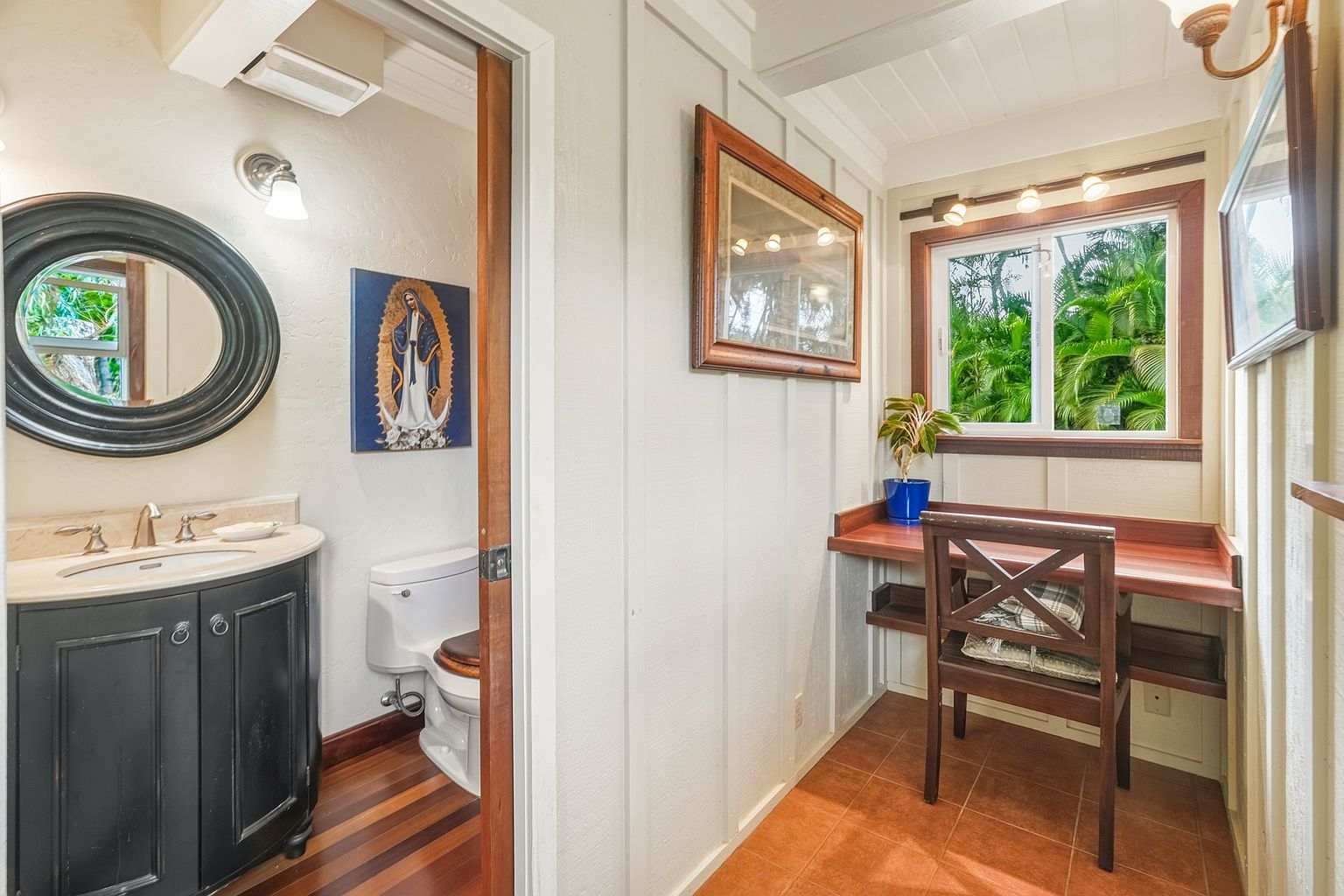 This image showcases a charming guest bathroom with a unique layout. The bathroom features a dark vanity with a marble countertop and a round mirror, complemented by a toilet and a framed religious painting. Adjacent to the bathroom is a small office nook with a wooden desk, chair, and a window offering natural light, creating a functional and aesthetically pleasing space.