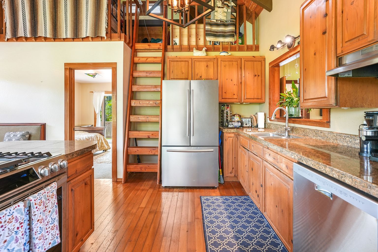 This is an interior shot of a rustic kitchen featuring wooden cabinetry, granite countertops, and stainless steel appliances. A unique wooden staircase leads to a loft area above the kitchen. The hardwood floors and a blue patterned rug add warmth and character to the space, creating a cozy and inviting atmosphere.