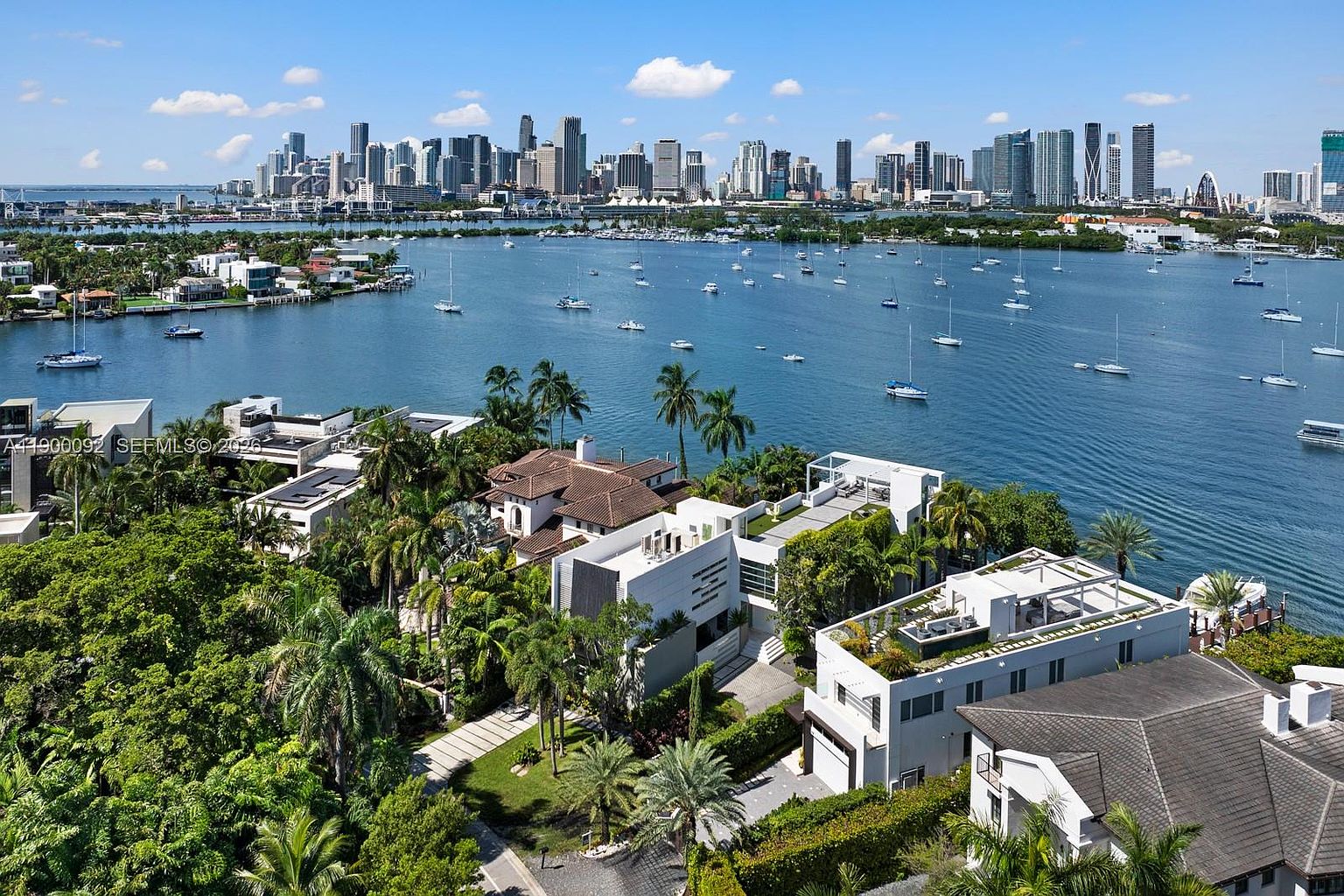 This high-angle aerial view captures a luxurious waterfront estate nestled among lush tropical landscaping, featuring a mix of modern and traditional architectural styles. The property boasts multiple levels, flat rooflines, and expansive terraces that overlook a serene bay filled with sailboats, with the impressive Miami skyline visible in the distance. The scene conveys an atmosphere of exclusivity and high-end coastal living, emphasizing the seamless integration between the private residence and the surrounding water.