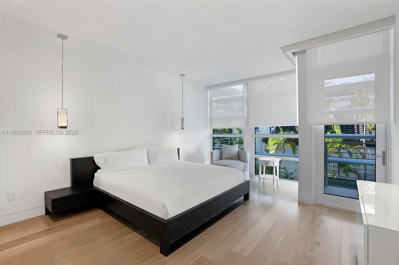 This bright and airy primary bedroom features a minimalist aesthetic with a dark wood platform bed frame and crisp white bedding. Large windows and a glass door provide ample natural light and access to a private balcony, while light wood flooring adds warmth to the space. Two sleek pendant lights hang above the bedside tables, contributing to the clean, contemporary design of the room.