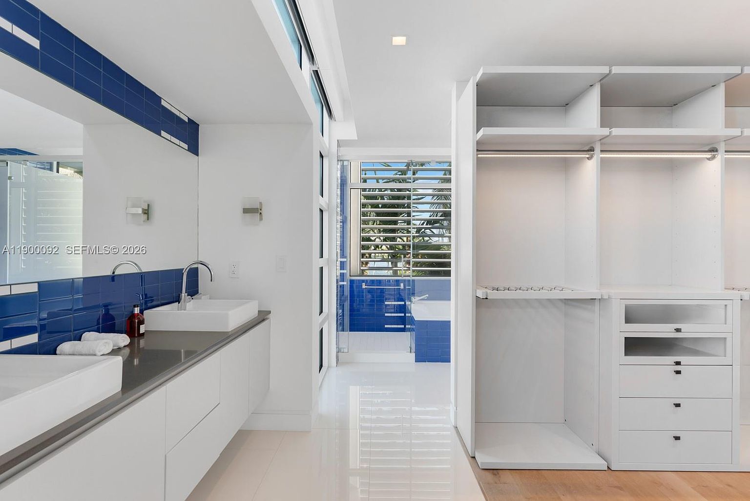 This modern primary bathroom features a sleek double vanity with white cabinetry and a dark countertop, complemented by a striking blue tiled wall. To the right, a large, open-concept walk-in closet with white shelving provides ample storage, while a glass-enclosed area in the background reveals a soaking tub. The space is bright and airy, with polished light-colored flooring and large windows that allow natural light to fill the room.
