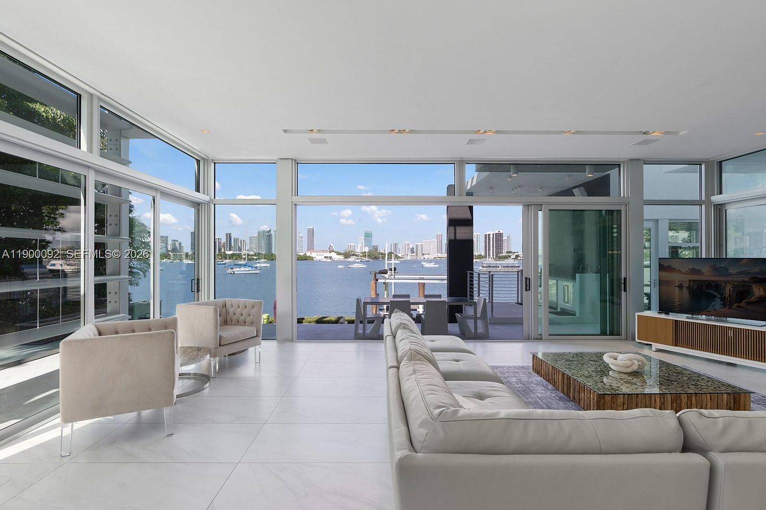 This expansive, modern living room features a neutral color palette with light-toned tile flooring and a large, plush sectional sofa. The space is defined by floor-to-ceiling glass walls that provide an unobstructed, panoramic view of the bay and city skyline. A low-profile media console and a glass-topped coffee table complete the minimalist aesthetic, creating a bright and airy atmosphere.
