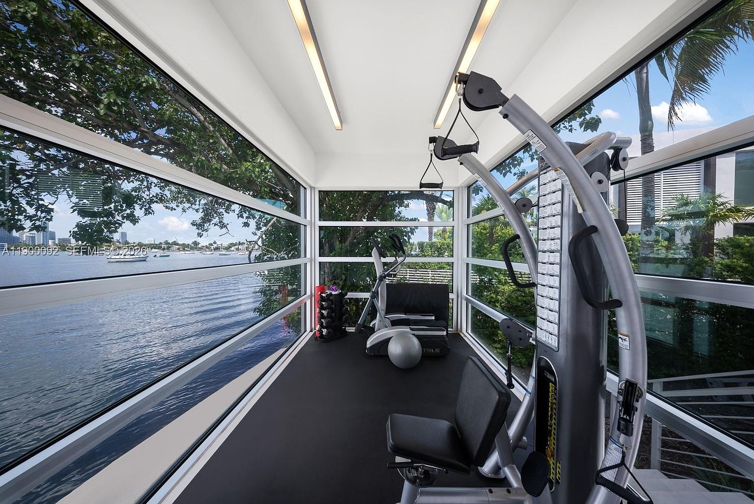 This home gym features a minimalist design with black rubber flooring and a multi-station strength training machine as the focal point. The space is enclosed by floor-to-ceiling windows on three sides, providing an immersive, panoramic view of the adjacent waterfront and lush greenery. The perspective is from inside the gym looking out, emphasizing the seamless connection between the workout area and the serene outdoor environment.