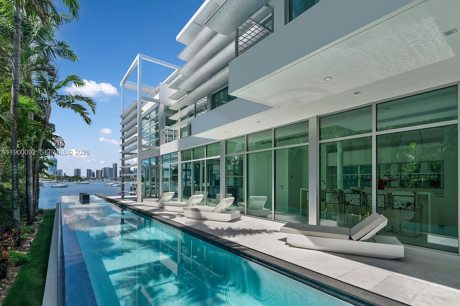 This modern waterfront estate features a sleek, rectangular lap pool that runs parallel to the home's expansive glass-walled facade. The outdoor living area is furnished with minimalist white lounge chairs, creating a serene space for relaxation against a backdrop of calm water and distant city skylines. The architecture emphasizes clean lines and a bright, contemporary aesthetic, seamlessly blending indoor and outdoor living.