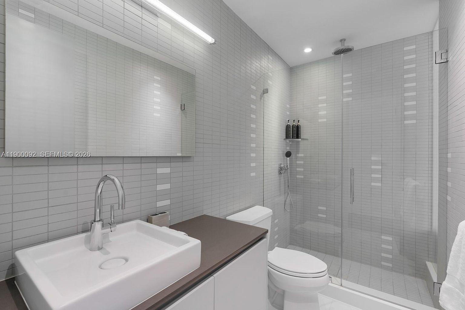 This modern, compact bathroom features a sleek, wall-mounted vanity with a dark countertop and a rectangular vessel sink. The walls are finished in a light-toned, grid-patterned tile that extends into the walk-in shower, which is enclosed by a frameless glass panel. A minimalist light fixture above the mirror and a rainfall showerhead contribute to the clean, contemporary aesthetic of the space.