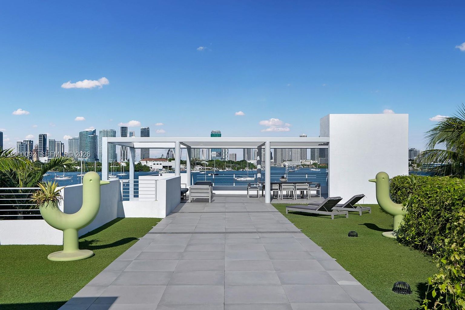 This expansive rooftop terrace offers a sophisticated outdoor living space with a paved central walkway flanked by artificial turf and whimsical, bird-shaped sculptural planters. The area features a modern pergola structure covering a dining set and lounge chairs, providing a perfect vantage point for panoramic views of the bay and the distant city skyline. The overall aesthetic is contemporary and resort-like, emphasizing an open-air lifestyle with a clear focus on the scenic waterfront backdrop.