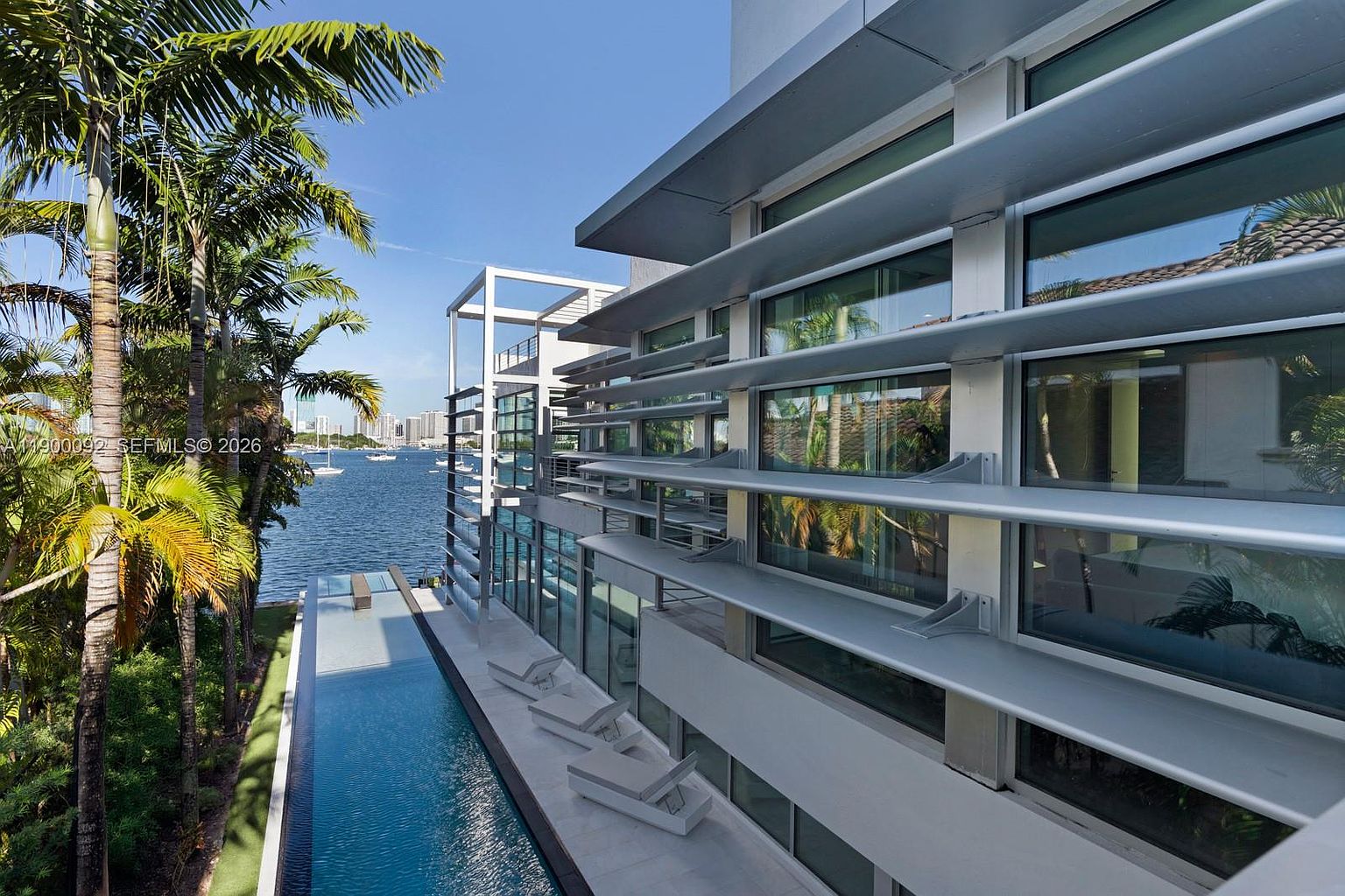 This modern waterfront property features a sleek, narrow lap pool running parallel to the building's side. The architecture is defined by horizontal metal louvers over large glass windows, creating a sophisticated, contemporary aesthetic. The perspective captures the length of the pool and the building's facade, with lush palm trees and a calm bay visible in the background.