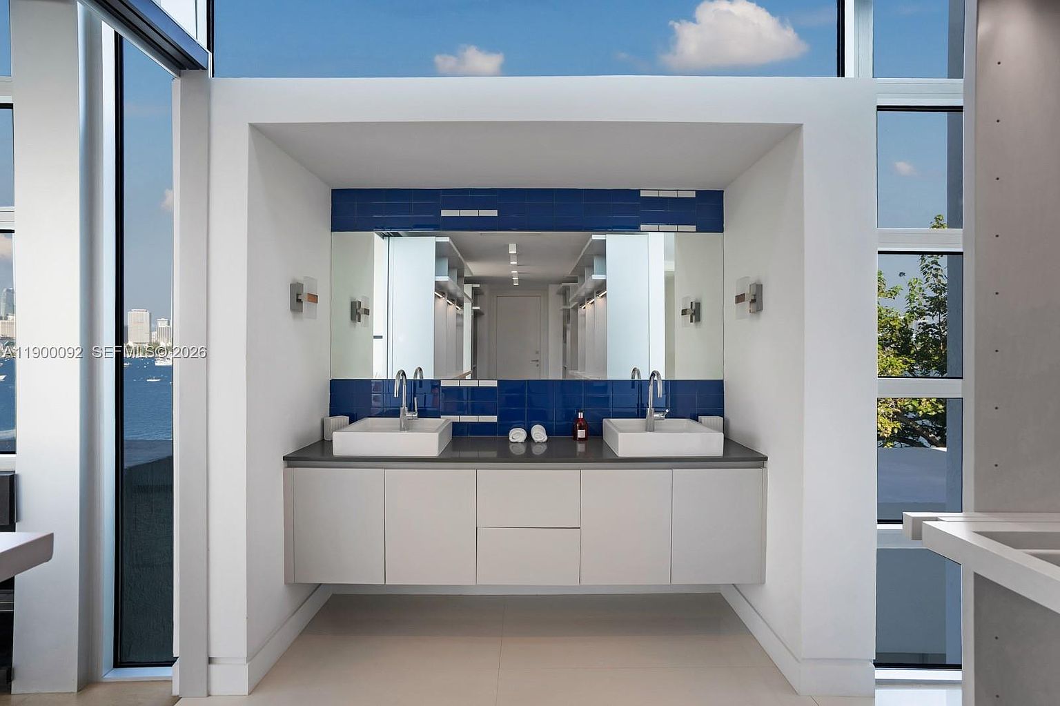 This modern primary bathroom features a double vanity with two square vessel sinks set against a striking blue tiled backsplash. The vanity is finished in a neutral tone with clean lines, and the space is framed by large windows on either side that offer expansive views of the water and city skyline. The overall aesthetic is sleek and contemporary, emphasizing natural light and a minimalist design.