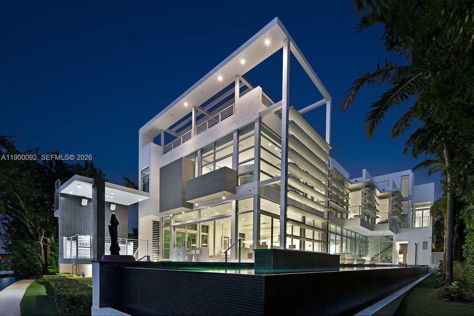 This striking contemporary residence is captured at twilight, showcasing a multi-level architectural design with extensive glass walls and clean, geometric lines. A sleek, dark-tiled infinity-edge pool stretches across the foreground, reflecting the illuminated interior and the structure's modern facade. The composition emphasizes the home's indoor-outdoor flow, featuring multiple balconies and a covered outdoor living area, all set against a clear night sky.