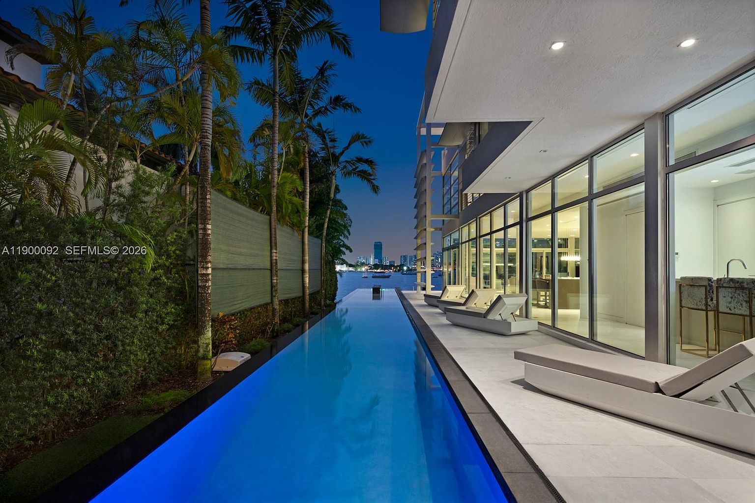 This stunning waterfront property features a sleek, narrow lap pool that stretches alongside a modern home with extensive glass walls. The patio area is furnished with contemporary lounge chairs, creating a serene outdoor living space that overlooks a calm bay at twilight. The composition emphasizes the seamless transition between the indoor living areas and the outdoor pool deck, highlighting the property's luxurious, resort-like atmosphere.