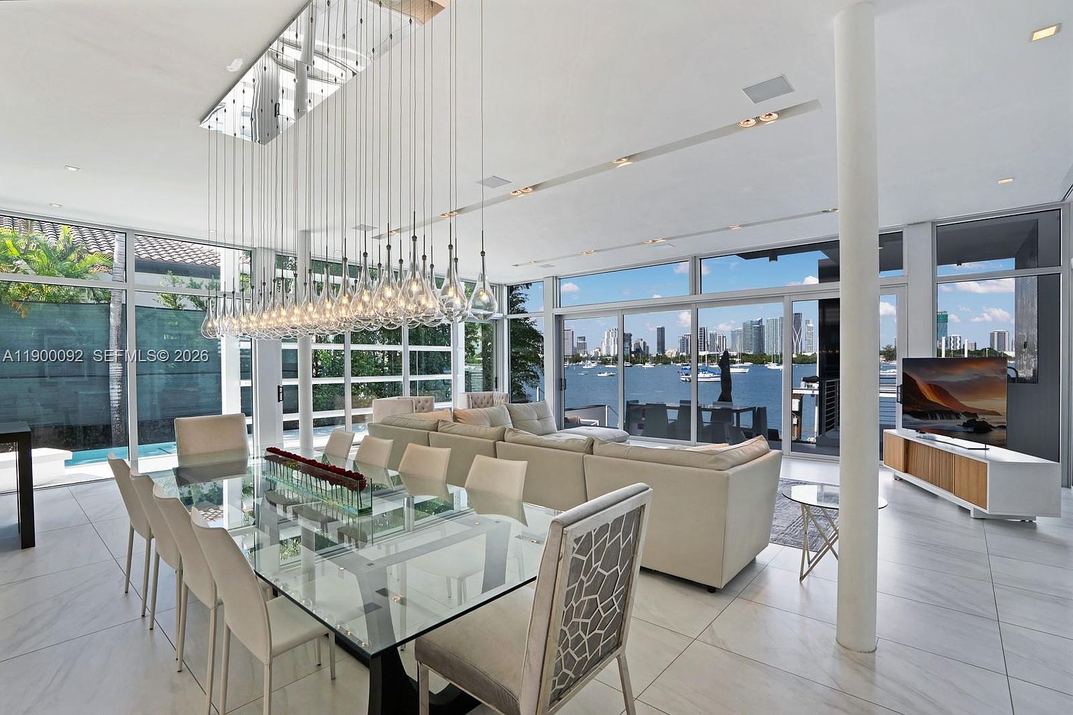 This open-concept living and dining area features a sleek glass-topped dining table surrounded by upholstered chairs, positioned beneath a dramatic, multi-pendant light fixture. The space is defined by large windows that offer expansive views of the waterfront and city skyline, creating a bright and airy atmosphere. A neutral-toned sectional sofa anchors the adjacent living space, while light-colored tile flooring runs throughout the room, emphasizing the modern, high-end aesthetic.