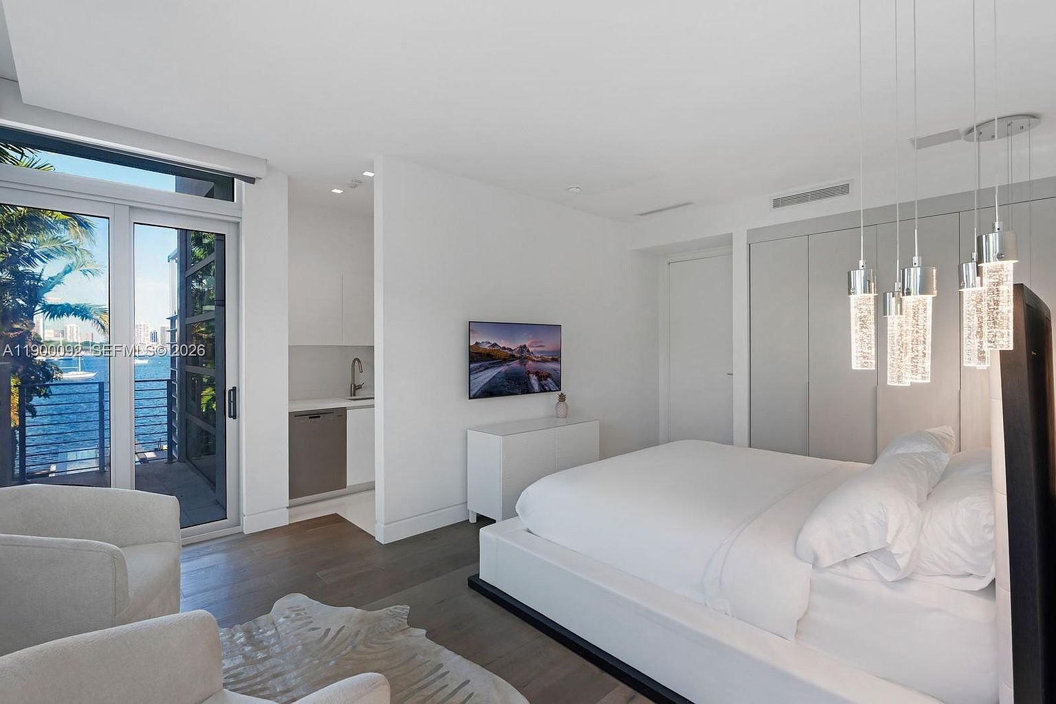 This modern primary bedroom features a minimalist aesthetic with a white platform bed, light wood flooring, and a neutral color palette. A small kitchenette area with a sink and dishwasher is tucked into an alcove, while floor-to-ceiling glass doors provide access to a balcony with water views. The room is accented by contemporary pendant lighting and a wall-mounted television above a sleek white console.