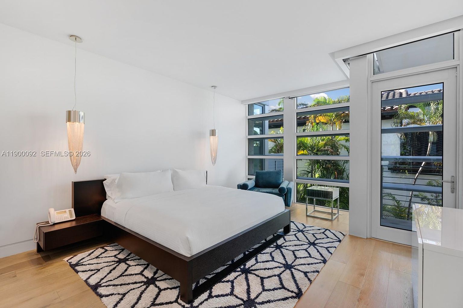 This modern primary bedroom features a dark wood bed frame centered against a crisp white wall, flanked by two sleek, hanging pendant lights. A large area rug with a geometric pattern anchors the space, while a blue armchair sits near the expansive windows that offer a view of lush tropical greenery outside. The room is bright and airy, with light wood flooring and a glass door providing access to an outdoor area.