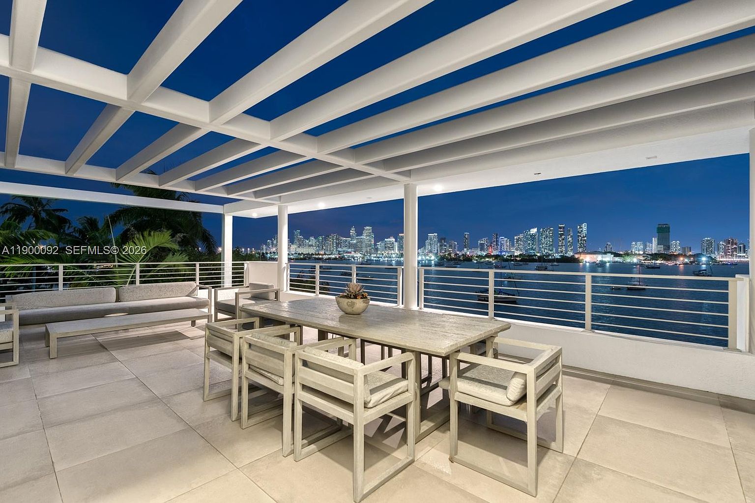 This expansive, covered balcony offers a sophisticated outdoor dining and lounge area with a panoramic view of a city skyline across the water at night. The space features a large, light-toned dining table with matching chairs and a long, cushioned sofa, all set beneath a modern white pergola structure. The clean, minimalist design and neutral tile flooring create a serene atmosphere, perfectly framing the vibrant urban lights in the distance.