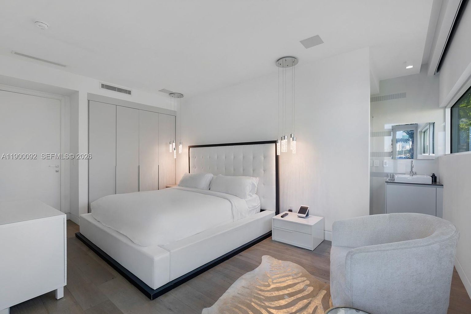 This modern bedroom features a plush white tufted headboard as the focal point, flanked by minimalist white nightstands and contemporary pendant lighting. A soft, patterned area rug sits beneath a curved armchair, while a built-in wardrobe with sleek, handleless doors provides ample storage. The room is bathed in bright, neutral tones, creating a serene and sophisticated atmosphere.
