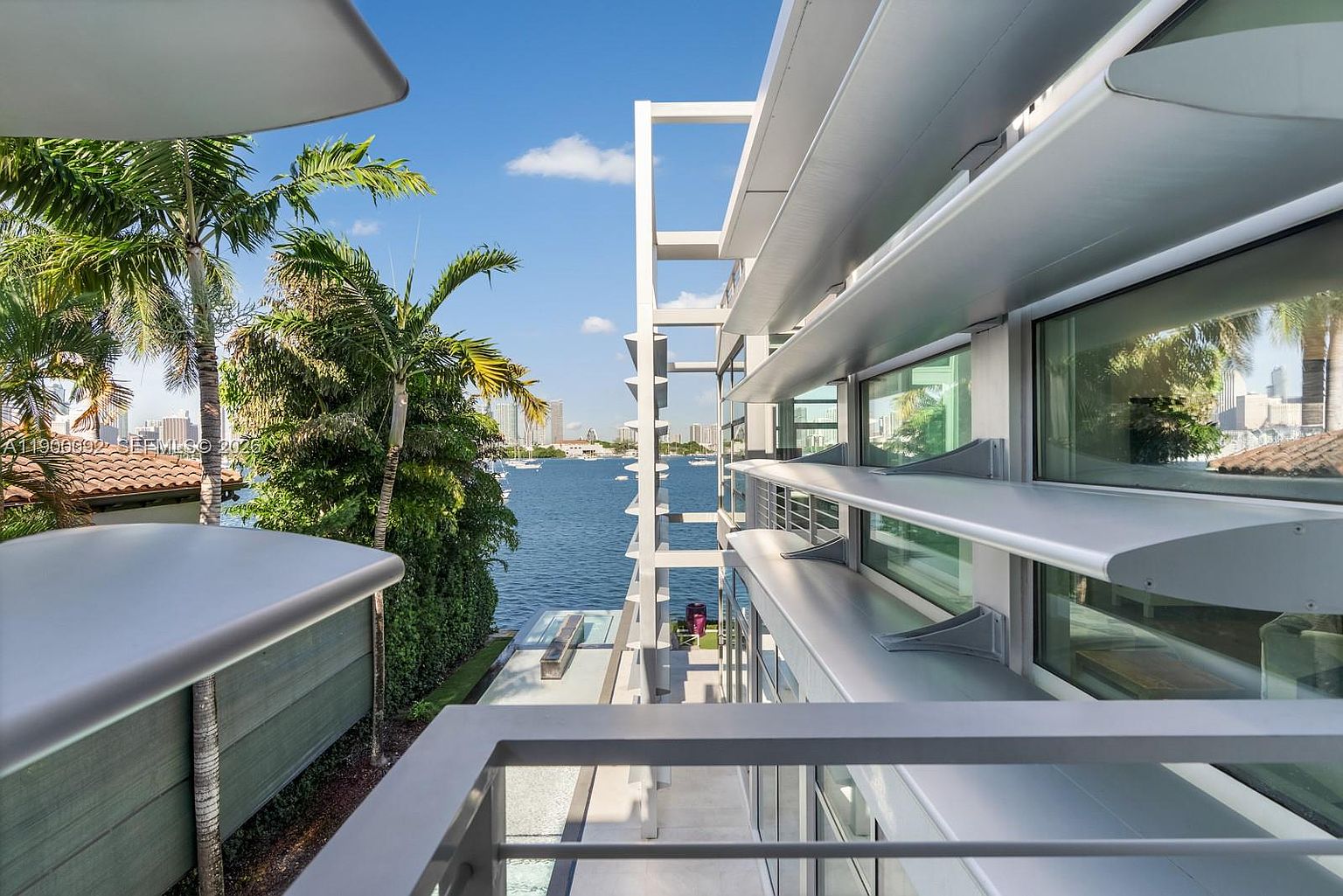 This perspective from a modern balcony overlooks a serene waterfront setting with a clear view of a distant city skyline. The architecture features sleek, horizontal metal louvers that provide shade and a contemporary aesthetic, while a glass railing offers an unobstructed view of the water below. Lush palm trees and greenery frame the scene, creating a tranquil and luxurious atmosphere.